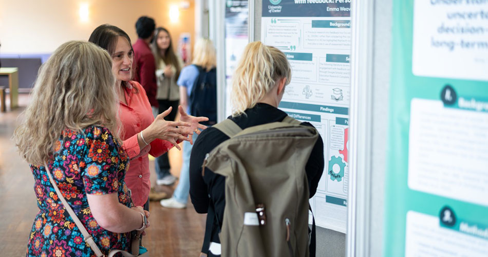 Two women standing indoors, engaged in conversation near a poster presentation.