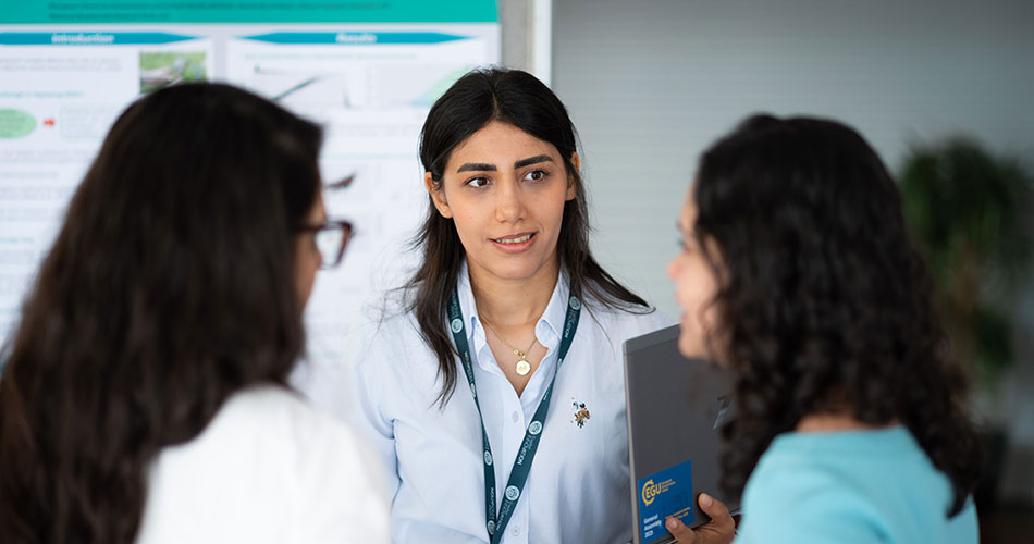 A woman wearing a lanyard and a light blue shirt engages in conversation with two other individuals at what appears to be an academic or professional event.
