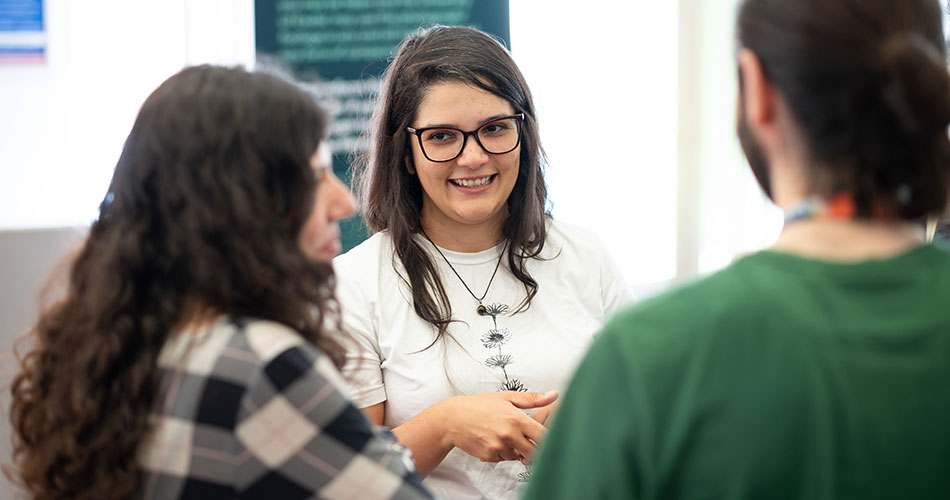 A woman with glasses and a white shirt smiles while talking to two other individuals in an indoor setting.