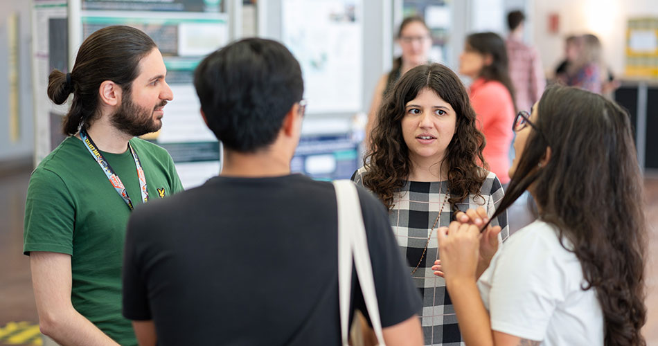 A group of four people engaged in conversation in a hallway, with one woman holding a pen and gesturing as she speaks.