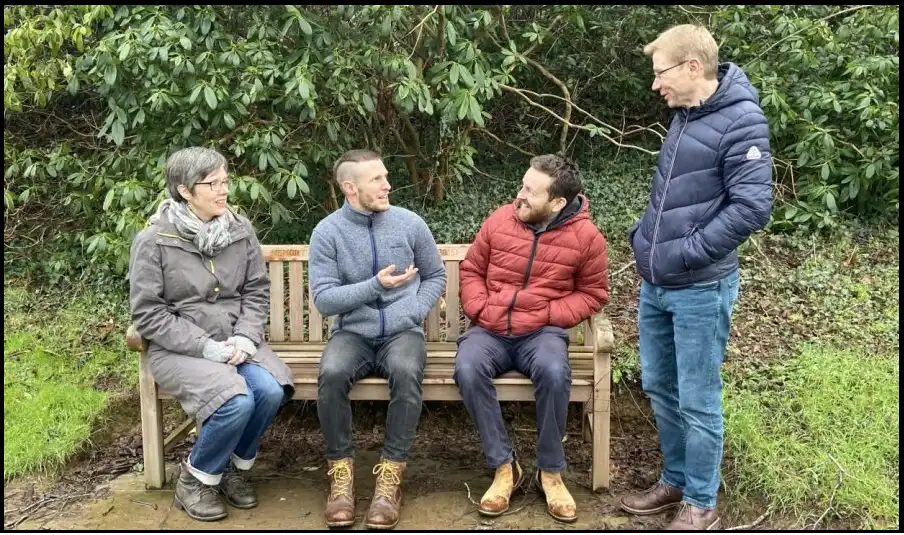 Four people in winter coats talking animatedly. Three are sat on a bench and the fourth stood next to the bench. There are trees and bushes behind them.